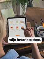 Een vrouw bekijkt haar favoriete Nederlandse ontbijtproducten en thee op een tablet. Artikel over Nederlandse ontbijtproducten eenvoudig te bestellen in het buitenland.